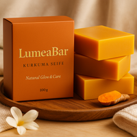Produktbild mit LumeaBar-Seifenbox, mehreren orangefarbenen Seifenstücken und frischer Kurkumawurzel auf einem Holztablett.